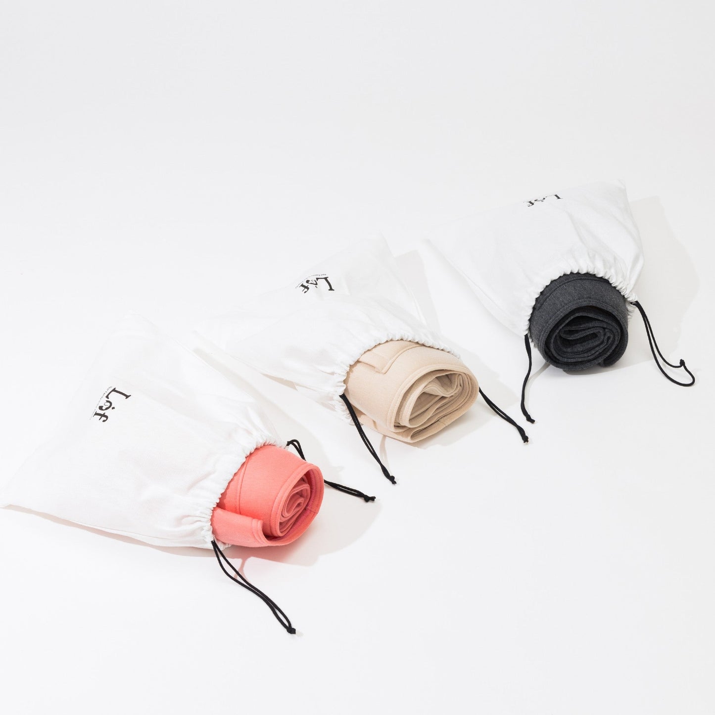 Three rolled-up towels in different colors (red, beige, black) on white bags with drawstrings.