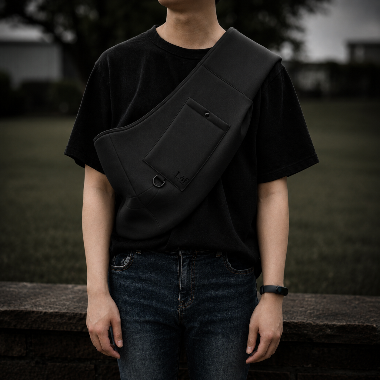A person wearing a black crossbody sling bag across their chest, paired with a black t-shirt and dark blue jeans, standing outdoors against a blurred background.